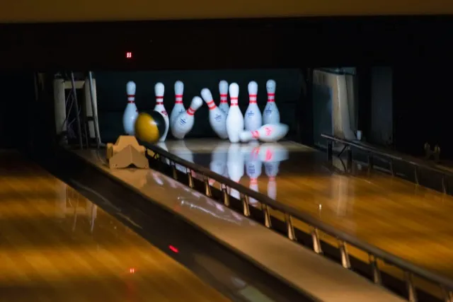 A black bowling ball strikes ten white pins with red necks on a glossy lane, several pins falling, perfect strike moment. A black bowling ball strikes ten white pins with red necks on a glossy lane, several pins falling, perfect strike moment.