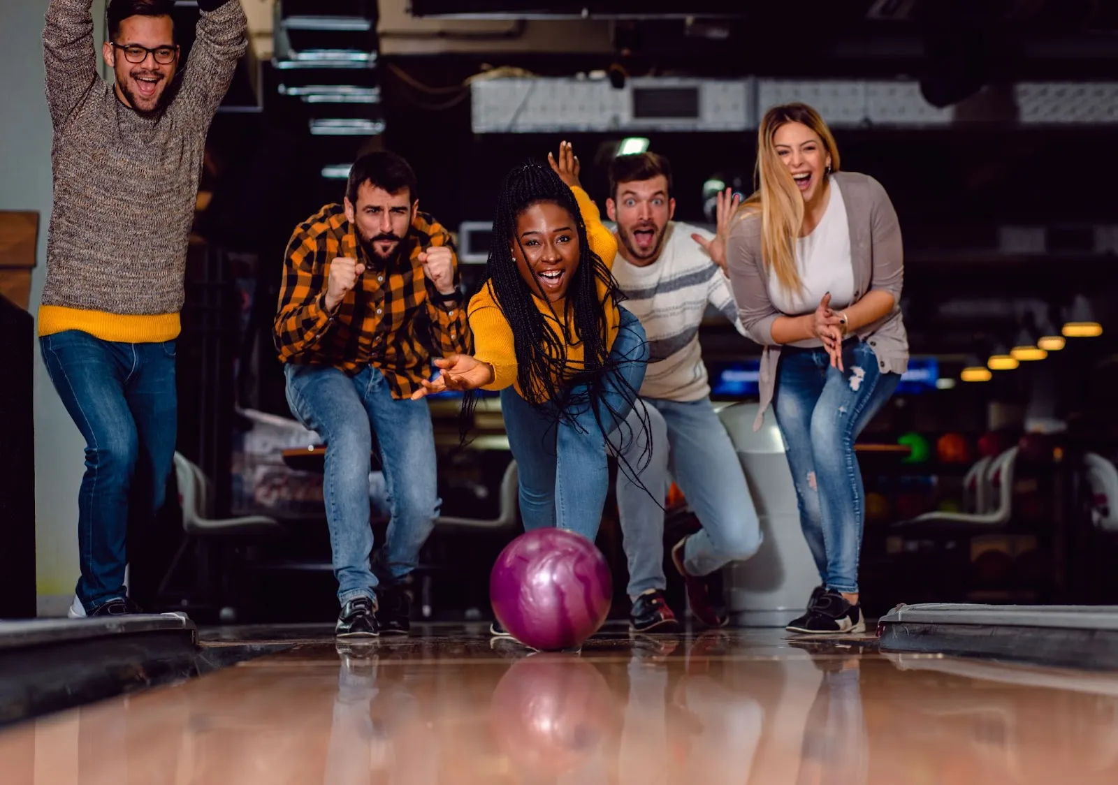 Friends having fun bowling together as a woman releases a purple ball down the lane while the group cheers excitedly. Friends having fun bowling together as a woman releases a purple ball down the lane while the group cheers excitedly.