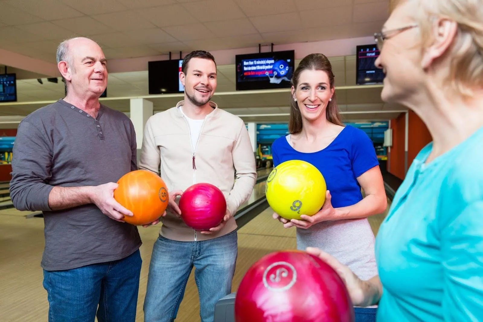 Three individuals stand in a bowling alley each holding a bowling ball preparing to take their turn