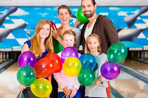 A joyful family at a bowling alley, each holding colorful balloons, celebrating a fun day together.