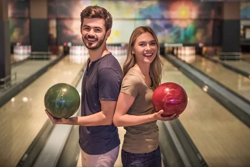 A man and woman stand in a bowling alley, each holding a bowling ball, ready to take their turn.