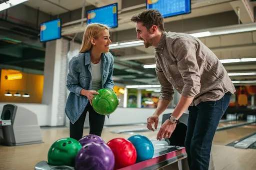 A man and woman are playing bowling, each poised to roll their balls towards the pins in a lively bowling alley.