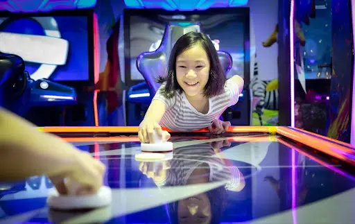 A young girl enthusiastically playing air hockey, focused on the game as she strikes the puck with determination.