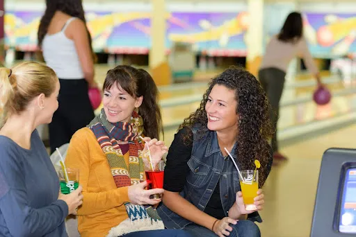 Three women sit together at a bowling alley, sipping drinks and enjoying their time.