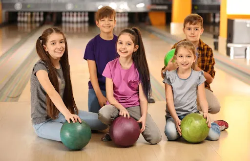 A group of children sitting on the floor, surrounded by colorful bowling balls, with the Skinny Dogz logo in the background.