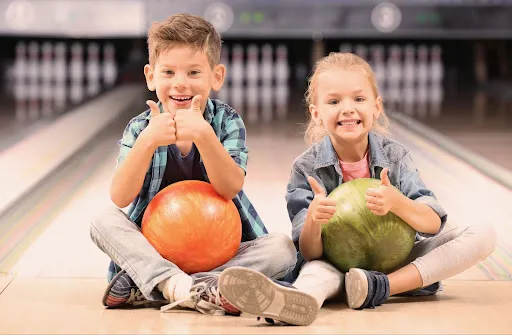 Two children on a bowling lane, sitting comfortably with bowling balls around them, sharing a joyful experience.