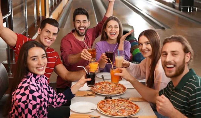 Friends-having-pizza-and-drinks-at-a-bowling-alley,-surrounded-by-bowling-balls-and-pins Friends having pizza and drinks at a bowling alley, surrounded by bowling balls and pins.
