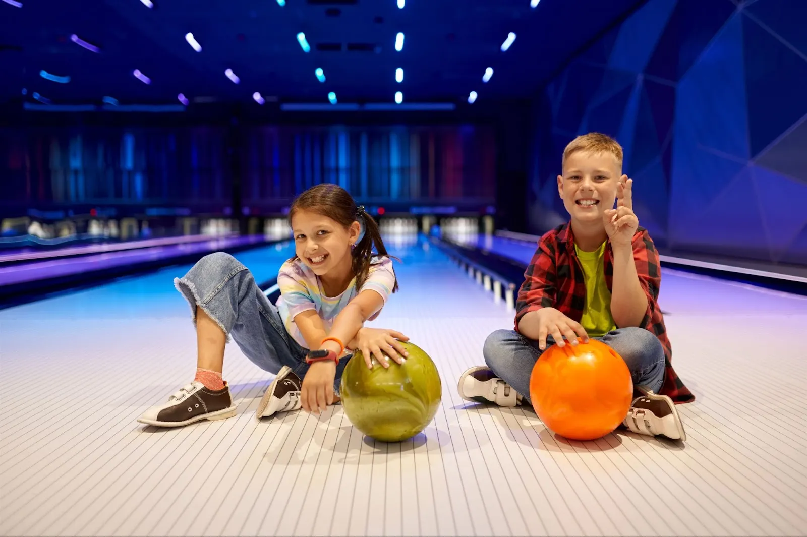 Two children seated on a bowling lane, surrounded by colorful bowling balls during a joyful family outing at the bowling alley.