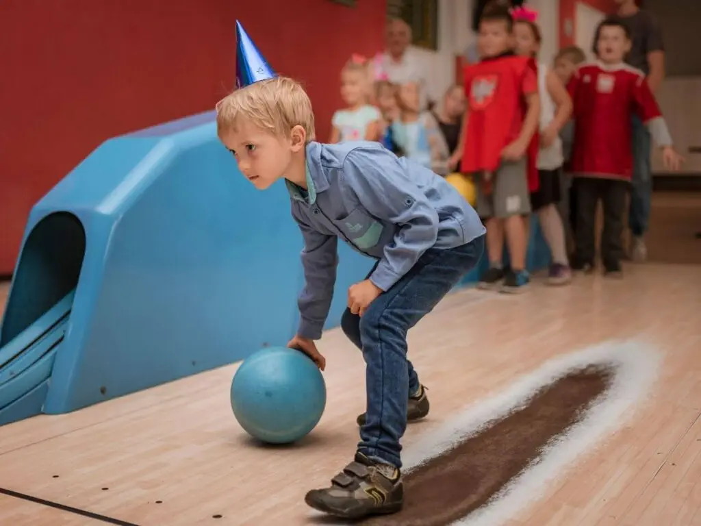A young boy in a birthday hat joyfully holds a bowling ball for his special day