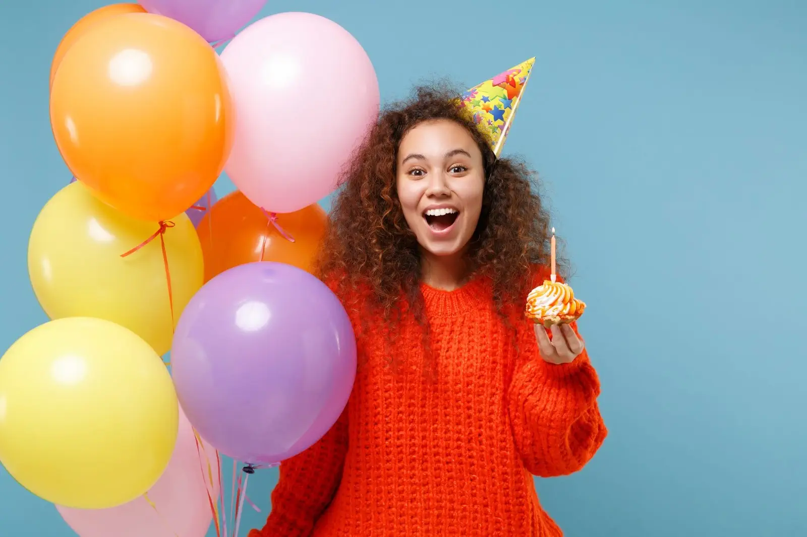 Smiling young woman in a party hat holding a cupcake with a lit candle, surrounded by colorful balloons.