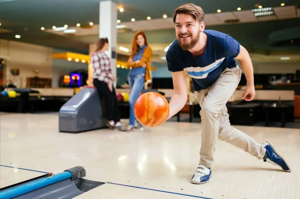 A man bowls with a ball while others watch and socialize in the background of a bowling alley