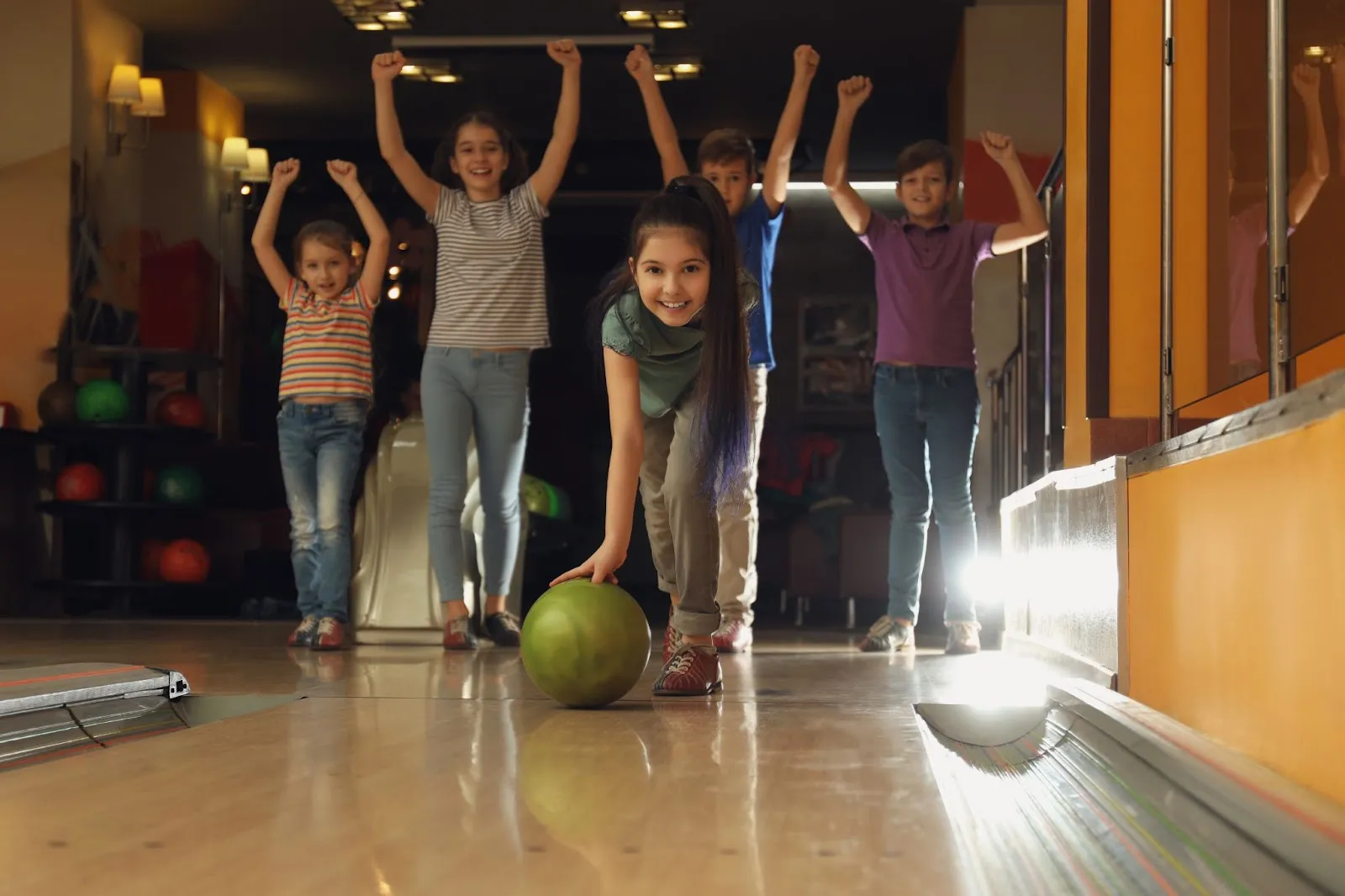 Children playing bowling, with one rolling a green ball and others cheering with raised arms.